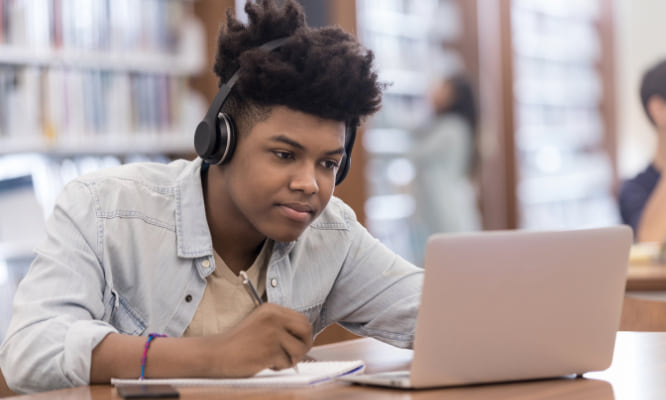 person in a library looking at a laptop and writing in a notebook